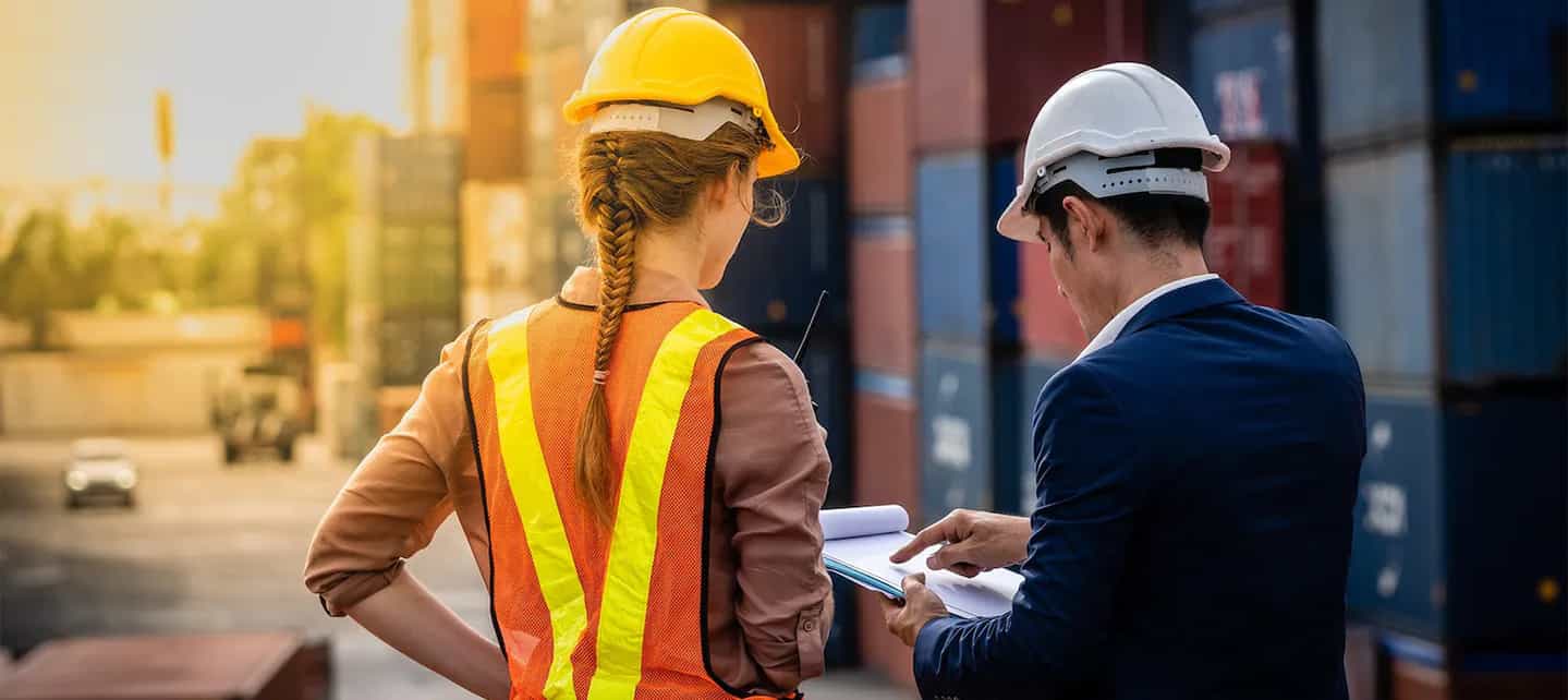 To personer i verneutstyr på en havn foran containere som ser på et dokument