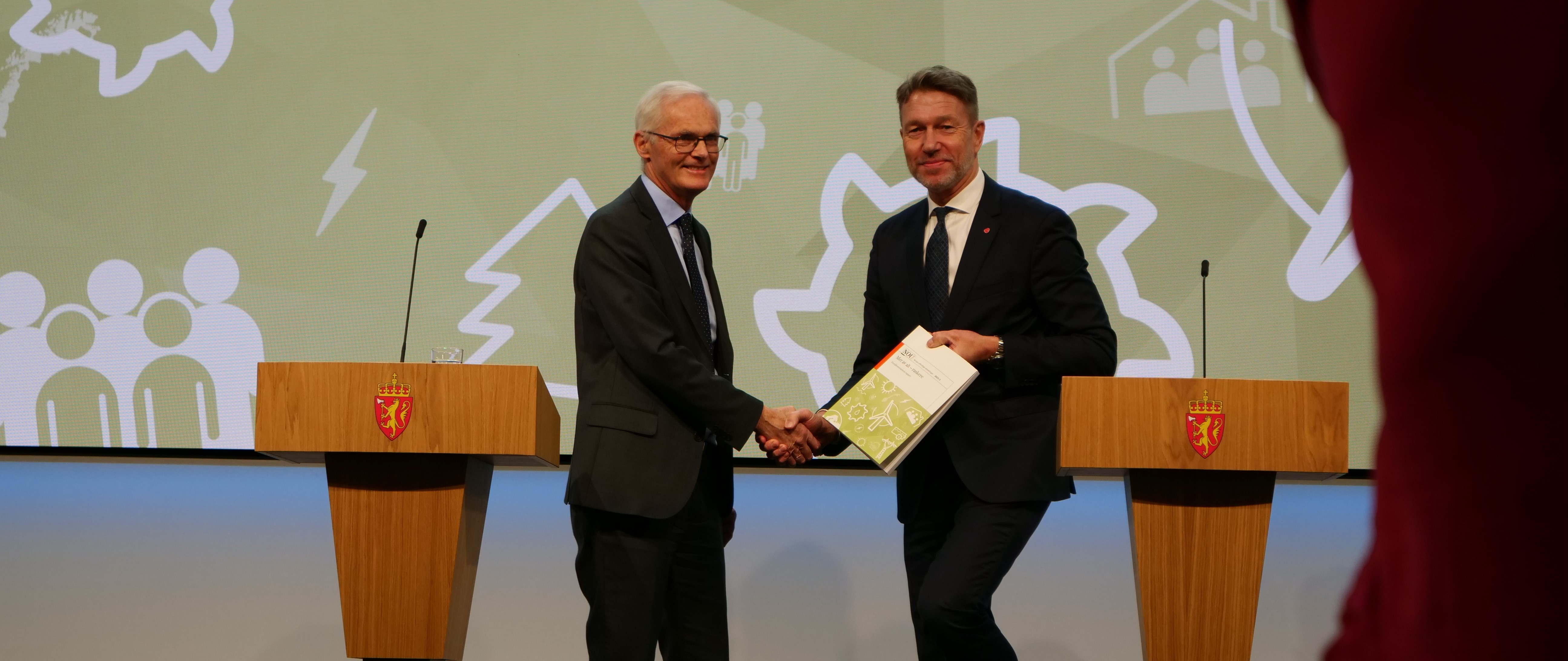 Energikommisjonens leder, Lars Sørgard og olje- og energiminister Terje Aasland stående på et podium med en rapport, og de tar hverandre i hånden. 